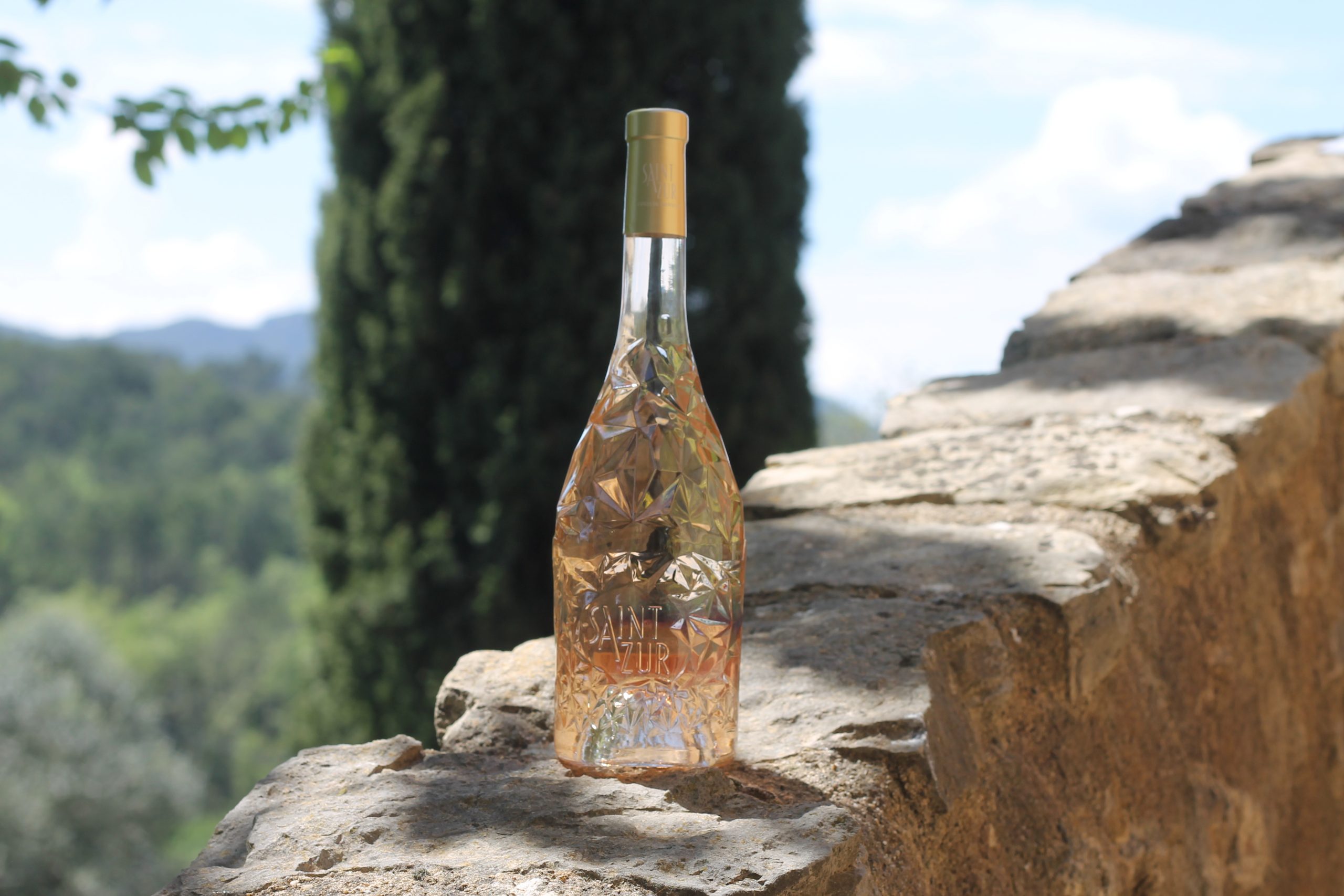 IMG_3369 Weinflasche SaintAzur mit diamantförmiger Glasoberfläche auf einer Steinmauer vor grüner Hügellandschaft.