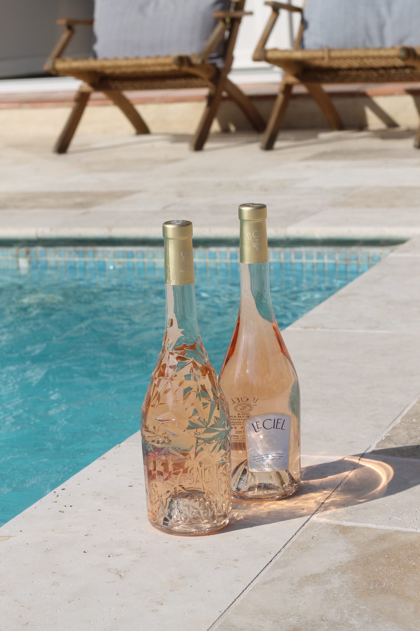 IMG_3377 Zwei Roséweinflaschen stehen am Rand eines Pools. Beide Flaschen haben goldene Verschlüsse. Im Hintergrund laden zwei Holzliegen mit Polstern zum Entspannen ein – ein sonniges, stilvolles Setting mit sommerlichem Flair.