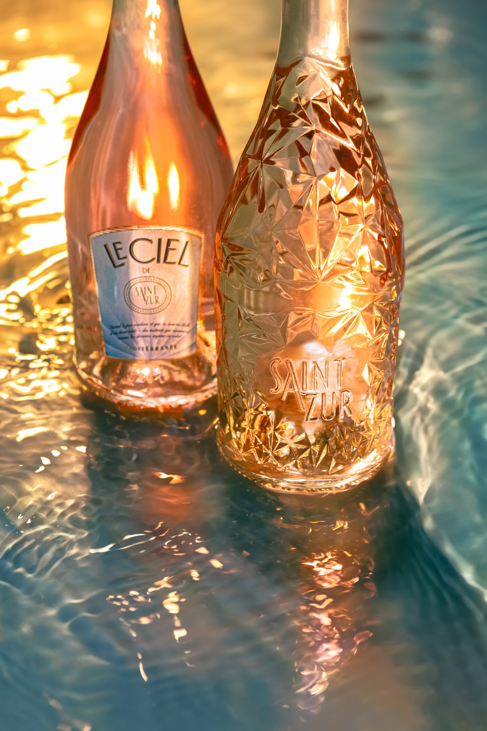 Zwei stilvolle Flaschen Roséwein, die teilweise in Wasser stehen. Das Licht reflektiert warm auf der Wasseroberfläche und den Flaschen, wodurch ein sommerliches, luxuriöses Ambiente entsteht.
